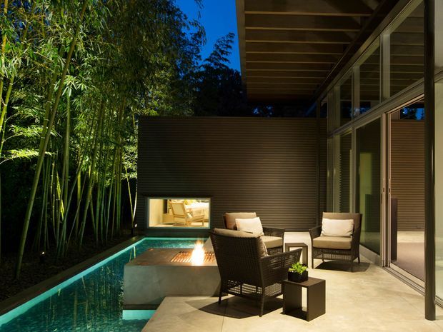 A sophisticated covered outdoor courtyard lounge at dusk, adjoining a glazed interior through large sliding doors. The space features dark horizontal wall cladding, exposed timber roof beams, a concrete fire feature beside a narrow reflective pool, woven outdoor armchairs, and dense bamboo planting along one edge.