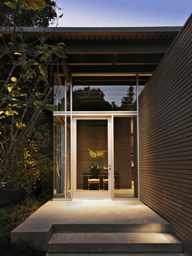 A modern residential entry viewed from outside, with broad concrete steps leading to a glazed front door and sidelights. Dark horizontal cladding, exposed roof framing, slim metal-framed glazing, and warm interior lighting create a refined, sheltered approach. Landscaping partially frames the entry.