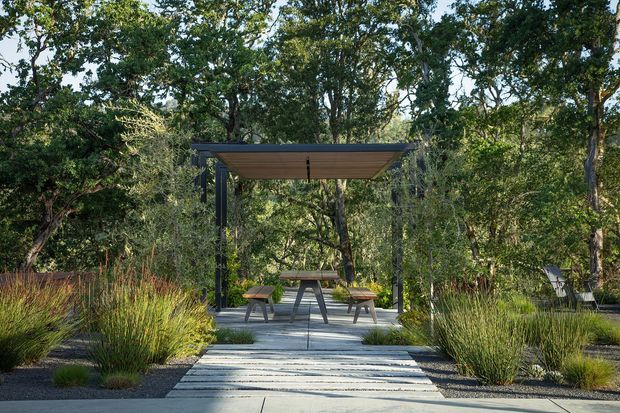 A landscaped outdoor garden setting with a contemporary open pavilion or pergola constructed from dark slender metal posts and a flat timber-lined roof. A picnic-style table and benches sit beneath it, reached by a broad pale paved path. Ornamental grasses, layered planting, mature trees, gravel beds, and a timber privacy fence surround the structure, creating a secluded natural retreat.