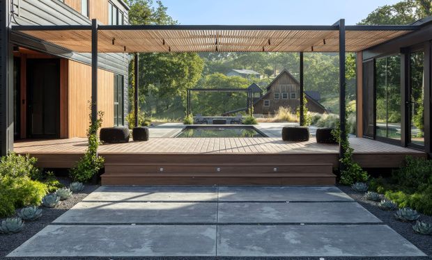 Contemporary outdoor living space viewed from a broad concrete walkway, with a raised timber deck, dark-framed glazing, timber-clad walls, and a slatted pergola creating a sheltered terrace. Minimal dark planters and low drought-tolerant landscaping frame the deck, while a green wooded setting and neighbouring pitched-roof building are visible beyond.