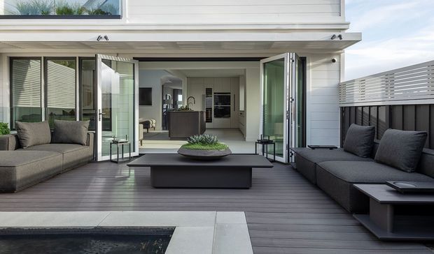 A contemporary outdoor entertaining area with dark timber decking, built-in grey seating, a low black coffee table, and a partially visible swimming pool in the foreground. Large bi-fold glass doors open into a bright interior living space, while white horizontal cladding and a slatted privacy screen frame the terrace.