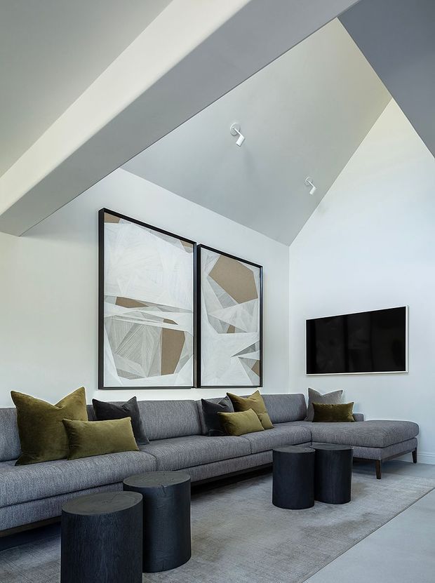 A bright, contemporary living room with a large grey sectional sofa, olive and charcoal cushions, black cylindrical coffee tables, and a pale area rug. White walls and a sharply angled vaulted ceiling create a spacious architectural backdrop, with two abstract framed artworks and a wall-mounted television.