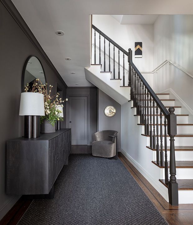 Refined residential entry hall with a staircase rising along the right side. The space features dark charcoal wall panelling and a matching built-in console, a large arched mirror, sculptural lamp and floral arrangement, grey upholstered armchair, patterned dark carpet, and a contrasting white stair enclosure with dark turned balusters and timber stair treads. Decorative wall mouldings and recessed ceiling lights add a tailored traditional character.