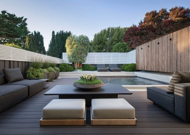 A refined private courtyard with a dark timber outdoor deck, low contemporary lounge seating, a broad coffee table, and a sculptural planter. The space opens toward a shallow rectangular pool or water feature, framed by layered clipped greenery, mature trees, horizontal screening, and a tall vertical timber privacy wall. The composition is symmetrical and secluded, with a calm, modern resort-like atmosphere.