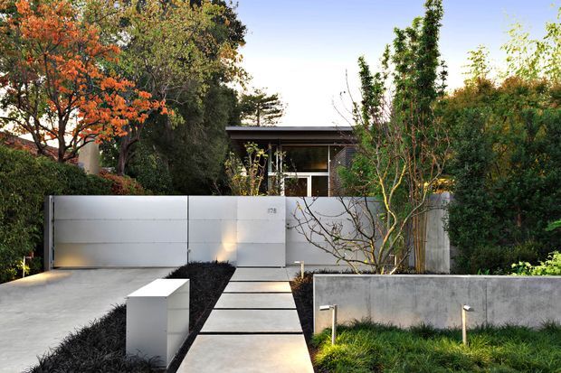 A contemporary single-storey residence viewed from the street, set behind a minimalist landscaped front garden. The approach features broad concrete paving, a central stepping-stone path, a smooth light-grey rendered or concrete boundary wall and gate, dark planting beds, integrated low garden lights, and dense established greenery with an ornamental tree. The house has a low, flat roof and dark-framed glazing partially screened by vegetation.