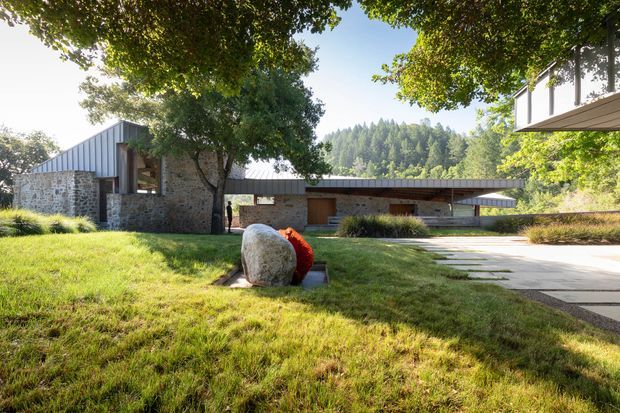 A low-slung modern residence set within a landscaped garden, featuring long horizontal volumes, dark metal pitched and skillion roof forms, extensive rough stone masonry walls, timber entry elements, and a paved driveway or walkway. Mature trees and a wooded hillside surround the house, while a sculptural boulder sits in the foreground.