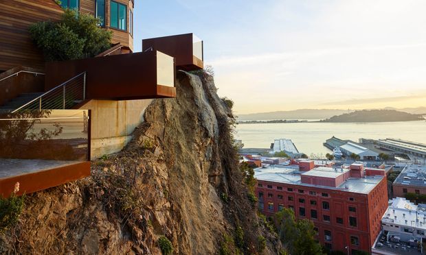 A contemporary multi-level building dramatically cantilevered over a steep rocky cliff, with warm timber cladding, dark metal volumes, exposed concrete, and glass windows. The view overlooks a marina, waterfront buildings, and a broad bay with hills in the distance.