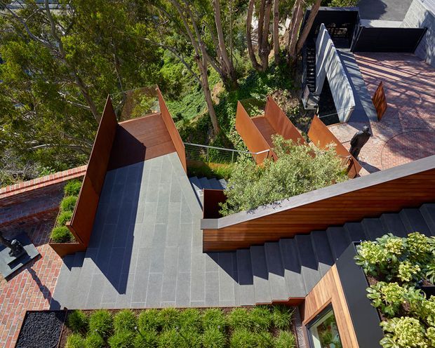 A high-angle exterior view of a contemporary multi-level residential property integrated into a steep, wooded site. Broad dark grey tiled or paved terraces and stairways connect angular rust-coloured metal-clad forms, warm timber-clad walls, planted roof sections, and a brick-paved courtyard. The composition emphasizes stepped circulation, geometric roof planes, and landscaped integration with mature trees.