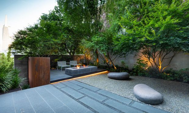 A contemporary landscaped courtyard and outdoor lounge with large grey paving slabs, inset rectangular stone detailing, a fine gravel garden, sculptural smooth boulders, dense mature planting, and a raised rectangular fire feature surrounded by seating. Rust-toned metal screening and warm integrated landscape lighting create a refined, secluded atmosphere. A slender building form is partially visible beyond the trees.