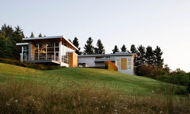 A low-slung modern house set on a gently sloping grassy site, with extensive glazing, projecting flat rooflines, horizontal weatherboard-style cladding, and a raised deck or balcony. Mature evergreen trees surround the property.