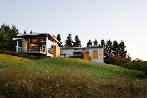 Exterior view of a low-slung modern house on a gently sloping grassy site. The home has strong horizontal lines, flat or very low-pitched roof forms, extensive floor-to-ceiling glazing, timber-toned wall panels, and a covered terrace or balcony. Mature trees surround the property.