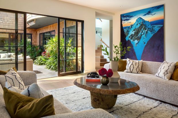 Bright, refined living room with a large textured sectional sofa, sculptural stone coffee table, neutral rug, and bold mountain artwork. Black-framed glazed doors open to a lush courtyard, while a timber-lined exterior area and stairway are visible beyond. The room combines warm natural materials, abundant daylight, and colourful botanical accents.