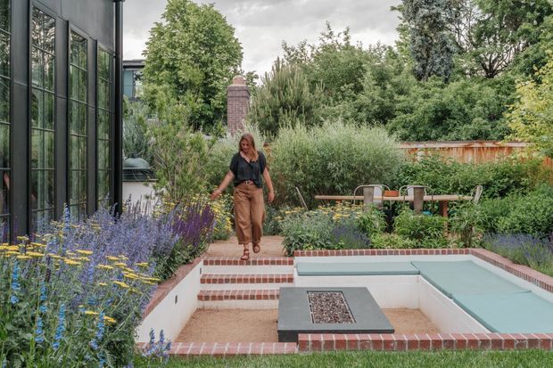 A landscaped outdoor courtyard garden beside a dark-framed glazed building, featuring a sunken rectangular seating area with pale built-in bench seating, brick edging and steps, a central fire-pit table, dense ornamental planting, and a dining table in the background. The setting has a lush, natural, layered garden design with a brick chimney or garden structure visible beyond.