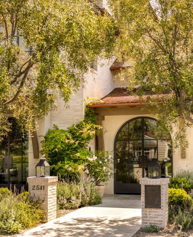 A warm, landscaped residential entrance viewed from outside. The facade features pale stucco or rendered walls, a red-brown tiled roof with layered rooflines, dark-framed glazing, and a large arched glass entry door. Brick-and-stone entry pillars with lanterns and the number 2541 flank a paved path, surrounded by mature trees and flowering garden planting.