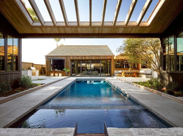 A refined courtyard-style outdoor pool area framed by dark timber-clad buildings and a covered timber structure with a glazed roof. The long rectangular swimming pool is bordered by pale stone paving, with a pavilion, outdoor seating, planting, and a built-in outdoor entertaining area visible beyond. Strong indoor-outdoor connection, abundant daylight, and warm timber detailing define the composition.