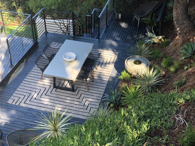 Elevated view of a landscaped outdoor dining deck surrounded by dense planting. The dark timber deck has metal railings, a rectangular dining table with chairs, a patterned outdoor rug, and a round fire bowl or water feature, creating a secluded garden entertaining area.