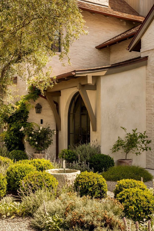 A warm, sunlit courtyard garden viewed toward the entrance of a stucco and pale brick residence. The facade features steeply pitched tiled roof sections, exposed timber eaves and brackets, and a tall arched glazed entry door. The landscaped foreground includes clipped box hedges, gravel, drought-tolerant planting, a small stone fountain or basin, and potted greenery.