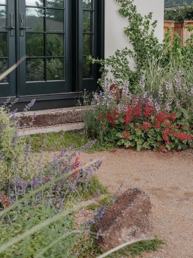 A close-up exterior view of a dark-painted, multi-pane glazed door and adjacent window set into a light rendered wall. Weathered timber steps lead to a gravel garden path bordered by abundant naturalistic planting, including grasses and flowering perennials. The scene has a refined yet informal cottage-garden character.