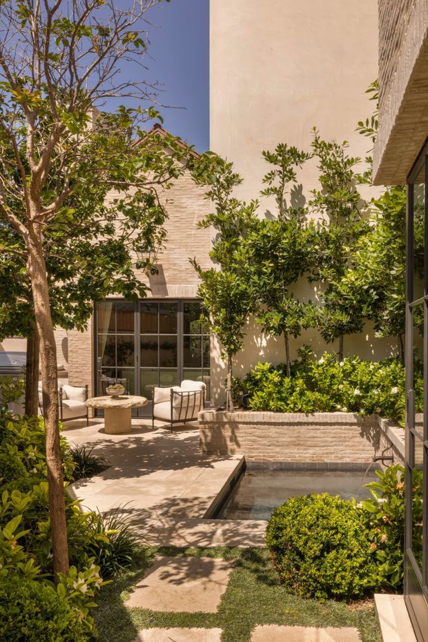 A sunlit landscaped courtyard with a small rectangular reflecting pool, paved stepping paths, mature trees, clipped shrubs, and lush planting. Warm pale masonry walls and textured brickwork enclose the space, with dark-framed glazed doors opening toward a seating area furnished with light lounge chairs and a round stone table. The courtyard has a calm, resort-like contemporary character.