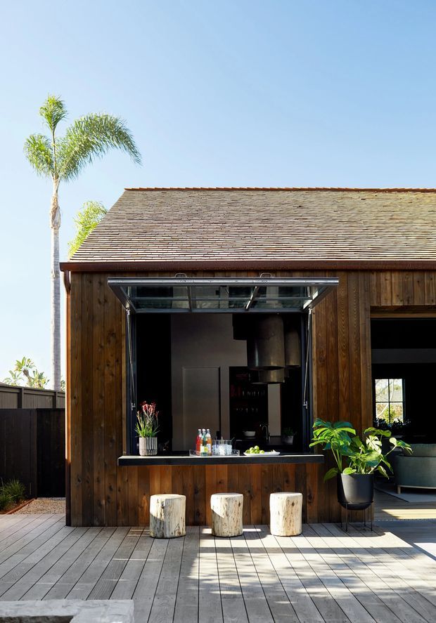 Exterior view of a compact timber-clad pavilion or outdoor entertaining building with a pitched shingle roof. A large open servery window with a dark counter creates an outdoor bar or kitchen connection, with three rustic log stools on a timber deck. Tropical planting and bright sunlight give the space a relaxed resort-like character.