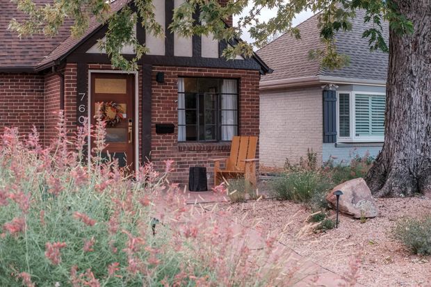 Exterior view of a compact brick house entry with a dark timber-framed front door, address numbers, divided-pane window, steep tiled roof, and a small paved path through established planting. An adjoining pale rendered wing with shutters is visible to the right, partly screened by a mature tree.