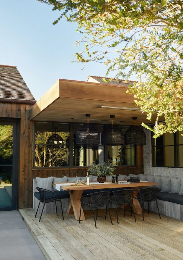 A sunlit outdoor dining terrace attached to a timber-clad home. The elevated timber deck features a long wood dining table, woven black dining chairs, built-in cushioned bench seating, and several large woven pendant lights beneath a broad timber-clad roof overhang. Glazed doors and windows connect the terrace to the interior, while mature tree branches frame the scene.