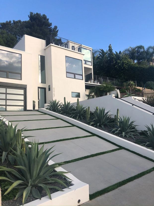 A modern multi-level home exterior with smooth pale stucco walls, dark-framed windows, a glazed garage door, rooftop and balcony terraces with metal railings, and a sloped driveway paved with large concrete slabs separated by narrow strips of grass. Agave and other drought-tolerant plantings line the driveway and retaining walls.