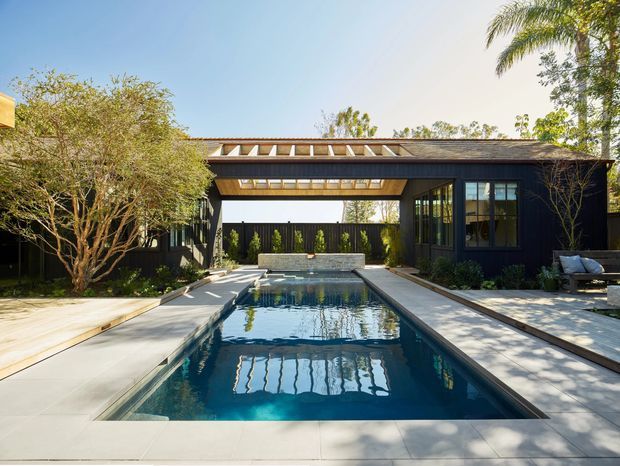 A refined contemporary courtyard centered on a long rectangular swimming pool. Dark-clad single-storey pavilion wings surround the pool, connected by a covered open-air passage with a timber-toned exposed roof structure and clerestory glazing. Pale stone or concrete paving extends around the pool, with mature trees and palms adding a landscaped, resort-like setting.