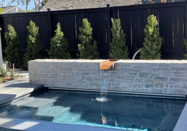 A compact outdoor swimming pool in a landscaped residential courtyard, bordered by a pale natural-stone masonry wall with a narrow water feature, dark timber fencing, and clipped screening plants. Paved pool surrounds and strong sunlight create a crisp contemporary garden setting.