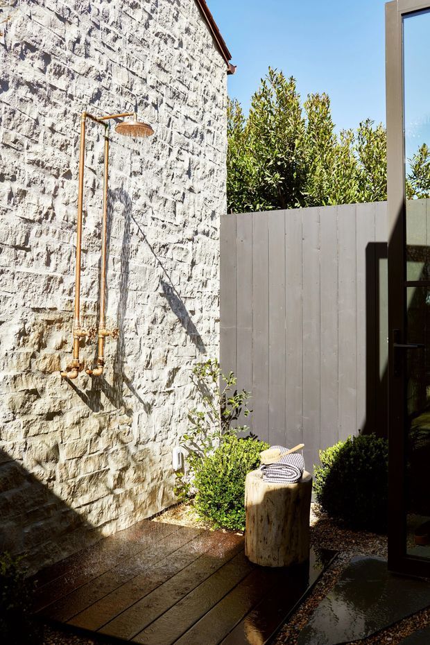 A sunlit outdoor shower courtyard with exposed rough stone masonry, copper plumbing and a round rainfall showerhead mounted to the wall. Dark timber decking, pale vertical fencing, clipped shrubs, a timber stool with folded towels, and a glazed dark-framed door create a private landscaped exterior space.