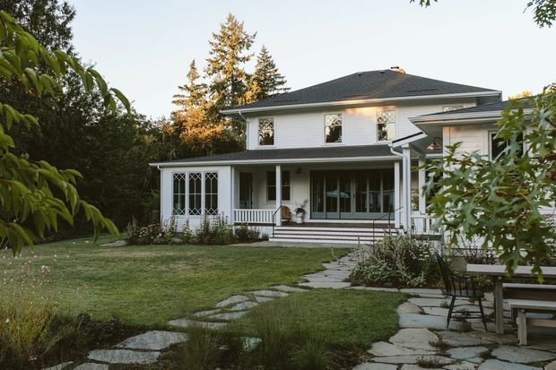 Exterior view of a two-storey white weatherboard house set within a landscaped garden. The home features a dark hipped roof, covered front veranda, decorative multi-pane glazing, white trim, and a stone-paved garden path leading toward the entrance.