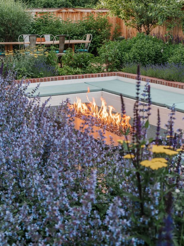 A landscaped backyard courtyard with a built-in outdoor fire pit surrounded by pale rendered walls and integrated bench seating. A brick coping border frames the fire feature, while lush flowering planting fills the foreground and edges. Behind it is a timber dining table with metal chairs, enclosed by a timber fence and dense greenery. The scene has a warm, intimate garden atmosphere.