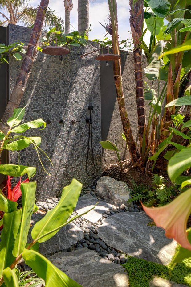 A lush tropical outdoor shower enclosed by dark textured pebble-stone walls, with black shower fixtures, a rain showerhead, and a natural stone stepping path surrounded by palms and broad-leaf plants. The setting feels like a private resort-style garden retreat.