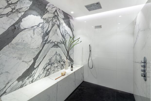 Luxurious contemporary walk-in shower featuring dramatic veined marble wall cladding, large white wall tiles, a white marble bench, dark flooring, and sleek chrome shower fixtures. Recessed ceiling lighting and ventilation provide a clean, refined atmosphere, with minimal decorative styling.