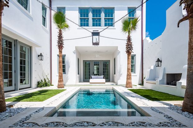 Bright white Mediterranean-style courtyard framed by multi-storey buildings, with tall palm trees, blue-trimmed windows, black lantern wall lights, a central rectangular swimming pool, and a built-in outdoor fireplace or seating alcove along the right wall.