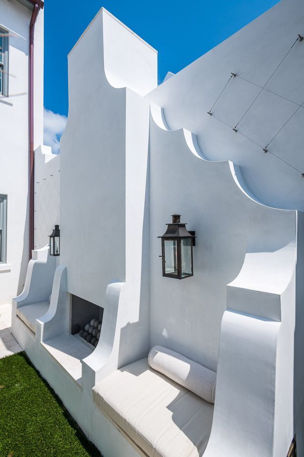 A bright exterior architectural detail featuring sculptural white stucco walls with repeated curved, scalloped forms and integrated built-in seating. Black lantern-style wall lights provide contrast, while a narrow strip of green lawn borders the space. The composition has a strongly Mediterranean-inspired, geometric and sculptural character under a clear blue sky.