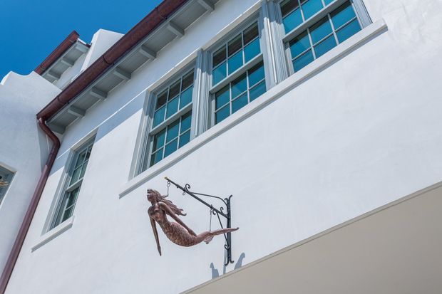 Upward-looking exterior view of a white rendered multi-storey building with tall, regularly divided windows, decorative vertical window surrounds, dark red-brown metal gutters and roof-edge details. A projecting wrought-metal bracket supports a mermaid-shaped sign or ornament on the facade. The image is dominated by the building envelope against a clear blue sky.
