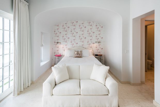Bright, softly styled bedroom with a white upholstered sofa at the foot of the bed. The room features an arched alcove framing the bed, pale pink flamingo-pattern wallpaper, white bedding and upholstery, sheer full-height curtains beside glazed doors, and a tiled floor. An open doorway on the right leads to a visible bathroom.