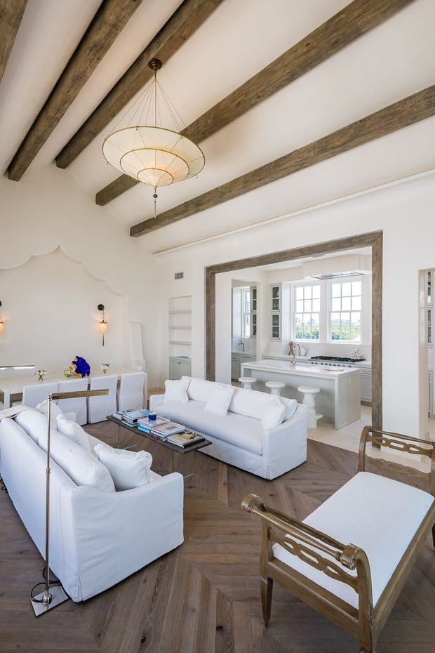 Bright, refined open-plan living room with white upholstered sofas and armchairs, a glass coffee table, white built-in cabinetry, and a visible white kitchen beyond a wide timber-trimmed opening. The room features a high white ceiling with exposed rustic timber beams, a sculptural pendant light, decorative wall moulding, and warm herringbone timber flooring.
