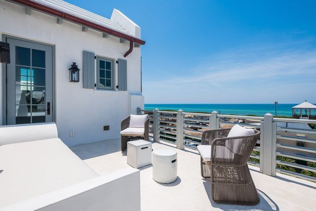 A bright rooftop or upper-level seaside terrace beside a white rendered building. The terrace features a white built-in daybed, outdoor lounge chairs, small white tables, and a horizontal metal balustrade, with expansive ocean views beyond. The building has blue-grey shutters, glazed doors and windows, exposed roof-edge elements, and red-brown guttering, creating a relaxed coastal setting.