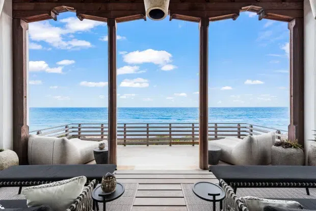 A refined open-air coastal lounge opening directly onto a timber deck and balustrade overlooking the ocean. The space features dark timber posts and ceiling beams, large open bays, pale upholstered lounge seating, dark woven daybeds, and a bright blue sea and sky beyond.