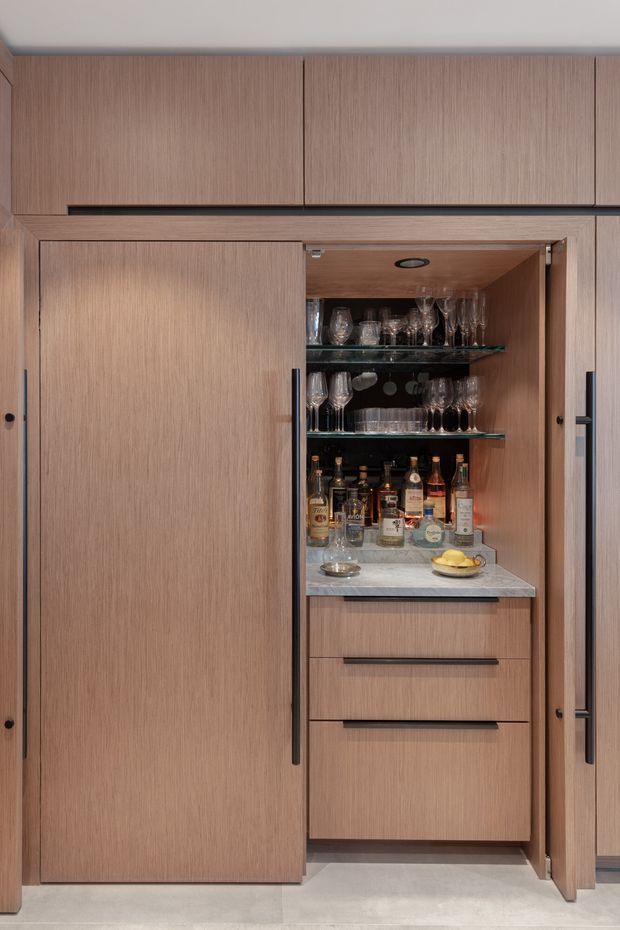Close-up of a built-in timber-toned bar cabinet with large sliding doors. The open section contains glass shelving, wine and cocktail glasses, liquor bottles, a pale stone benchtop, integrated lighting, and several drawers below. The surrounding cabinetry has slim dark handles and a warm, refined contemporary finish.