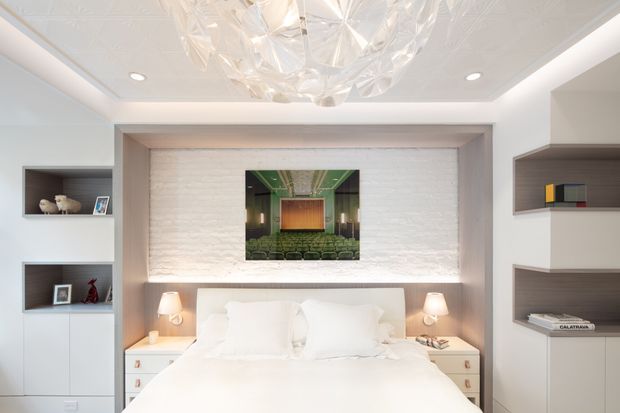 Bright, refined bedroom with a large upholstered bed, integrated bedside tables, and symmetrical built-in shelving. The bed wall is framed by a recessed architectural surround with a textured white feature wall and soft integrated lighting. A sculptural translucent chandelier, recessed ceiling lights, pale cabinetry, and curated decorative objects complete the polished contemporary interior.