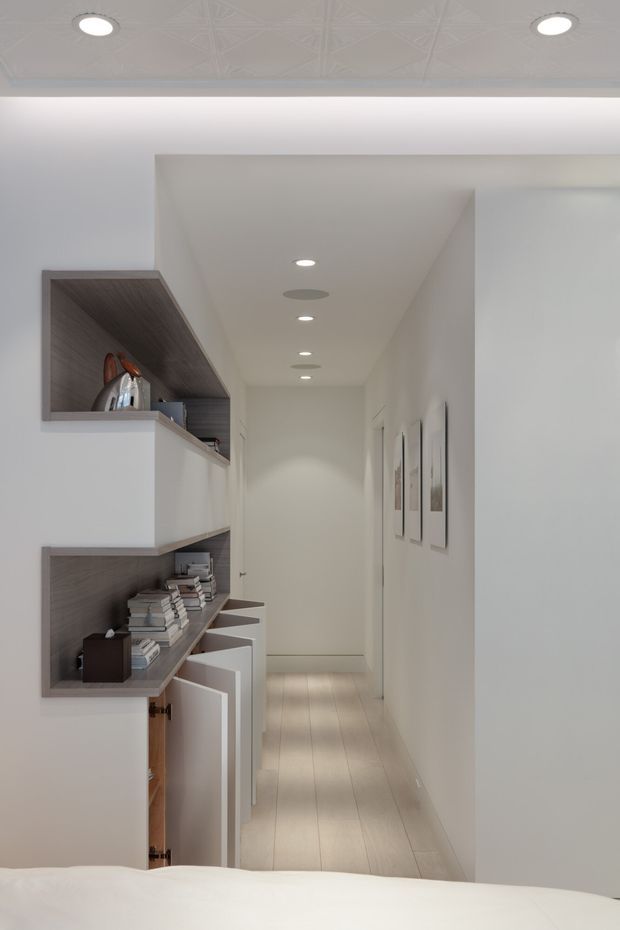 A bright, narrow residential hallway viewed from a bedroom, with white walls, pale timber flooring, recessed ceiling lights, and built-in open shelving and cabinetry along the left wall. The shelving displays books and decorative objects, while framed artwork lines the right wall. A patterned pressed-tin ceiling section is visible in the foreground, and the lower edge of a bed confirms the bedroom vantage point.