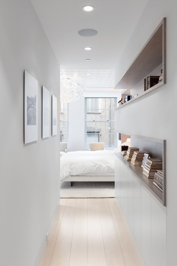 Bright minimalist bedroom viewed from a narrow hallway, featuring white walls and cabinetry, pale timber flooring, a white upholstered bed, recessed ceiling lights, framed monochrome artwork, and a built-in wood-lined shelving and display niche filled with books. Large windows bring in abundant natural light, creating a clean, airy contemporary interior.