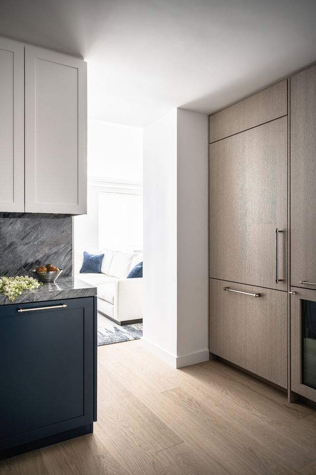 A bright, refined contemporary kitchen viewed toward an adjoining living room. The kitchen features dark navy-blue lower cabinetry with slim metal handles, pale grey-white upper cabinets, a dramatic dark veined stone splashback and benchtop, and a full-height bank of warm grey wood-grain cabinetry incorporating an oven. Light timber flooring, white walls, and natural daylight create a calm, cohesive space. A white sofa with blue cushions is visible beyond the kitchen opening.