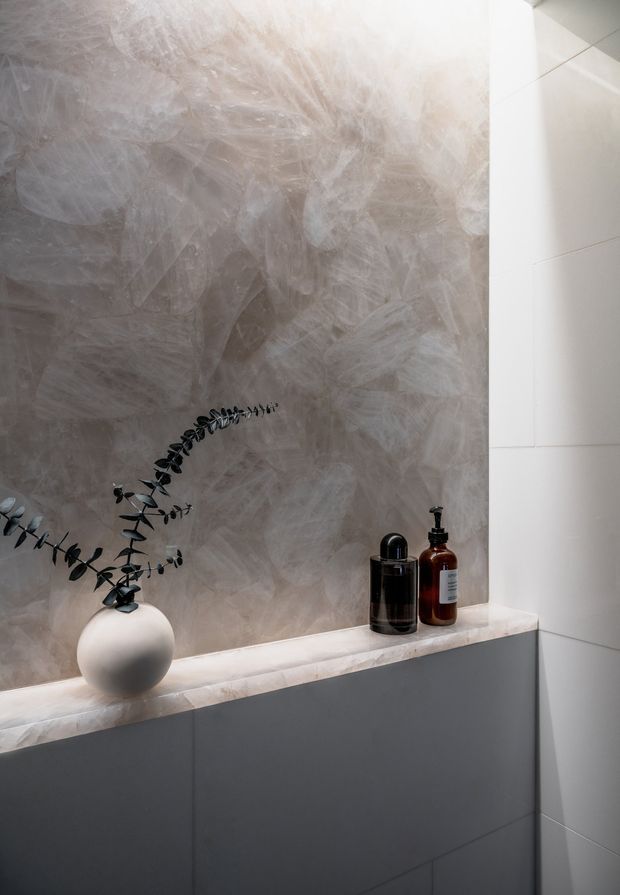 Close-up of a refined bathroom vanity or built-in cabinet. A grey wall has a layered, expressive plaster-like finish, meeting a pale stone or engineered-stone benchtop above dark grey flat-panel cabinetry. White large-format wall tiles frame the right side. Minimal decorative styling includes a round white vase with eucalyptus stems and two dark amber and black toiletry bottles.