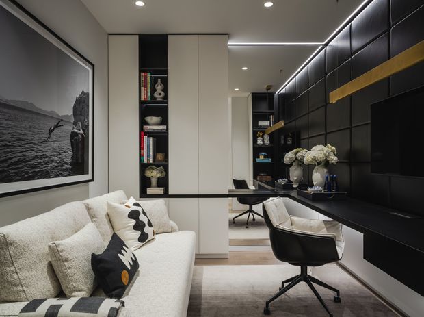 A refined contemporary living and work area with a light textured sofa in the foreground, a black feature wall with padded square panels and integrated cabinetry, a wall-mounted television, and a compact desk with upholstered swivel chairs. Full-height white built-in shelving and cabinetry provide storage, while linear LED lighting and recessed downlights create a polished, layered interior. A large monochrome artwork and neutral rug complete the sophisticated black, white, and grey palette.