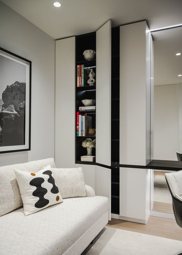 A refined contemporary living room corner with a pale upholstered sofa, sculptural patterned cushion, framed monochrome artwork, and a tall custom built-in storage wall. The joinery combines matte white doors with black open shelving and display niches, while a large mirror reflects part of the room. Recessed ceiling lighting and light timber flooring create a calm, polished interior.