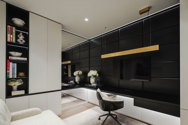 A sophisticated open-plan living and home office area with a dramatic black padded feature wall, integrated television, floating white cabinetry, and a long timber shelf. A compact desk and sculptural office chair sit beside the built-in joinery, while a white sofa and curated shelving occupy the foreground. The space uses concealed linear lighting, pale timber flooring, and a restrained monochromatic palette with warm timber accents.
